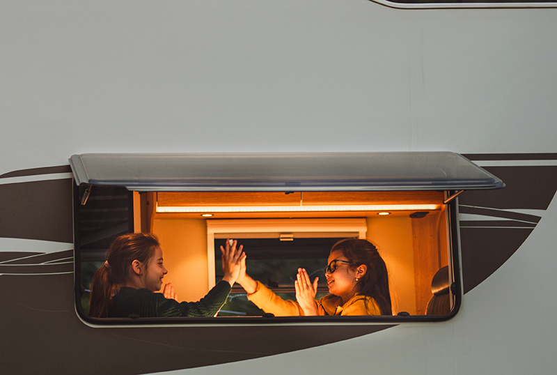 Photo prise de l'extérieur de deux fillettes jouant avec leurs mains à l'intérieur d'un camping-car éclairié.