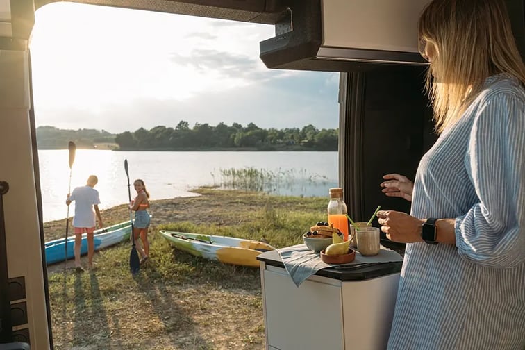 Une femme blonde vêtue d'une robe est à l'intérieur du fourgon et regarde vers l'extérieur. Deux adolescents préparent des kayak devant le lac.
