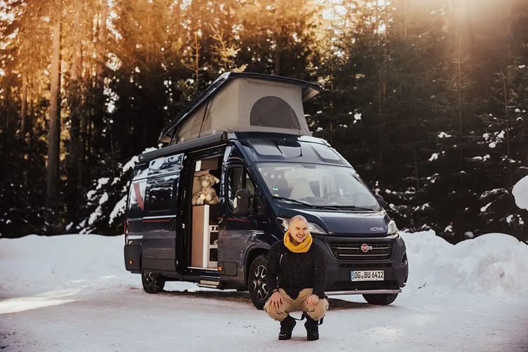 Un homme en tenue d'hiver, accroupi sur la neige devant son fourgon. Le fourgon est noir, le toit relevable et la porte latérale sont ouvertes. Dans le fond, une forêt enneigée.