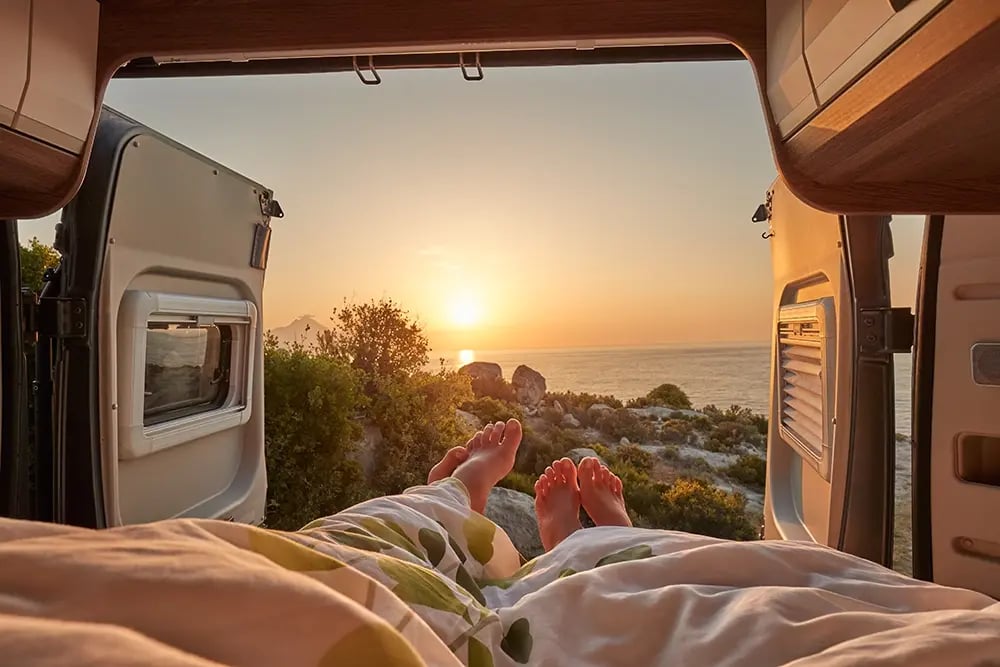 Photo prise depuis l'intérieur d'un fourgon aménagé. Les pieds de deux personnes dépassent de sous la couette, vue sur le soleil couchant sur la mer.