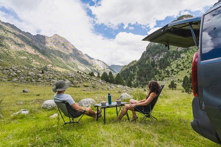 Un couple de vanlifer assis sur des chaises de camping avec une petite table, derrière leur van. Vue dégagée sur les montagnes verdoyantes.